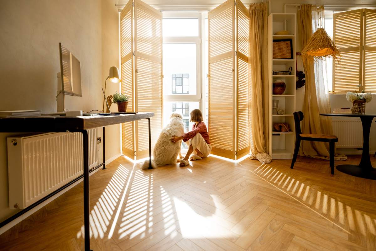 Young woman sits with her dog by the window blinds and looks away in cozy and sunny living room of modern apartment in beige tones. Quarantine, loneliness and life at home concept