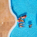 Aerial vertical view of group of women friends have fun together in summer holiday vacation at fibreglass pool resort with coloured inflatables lilos on the blue water