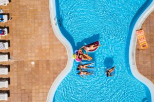 Aerial vertical view of group of women friends have fun together in summer holiday vacation at fibreglass pool resort with coloured inflatables lilos on the blue water